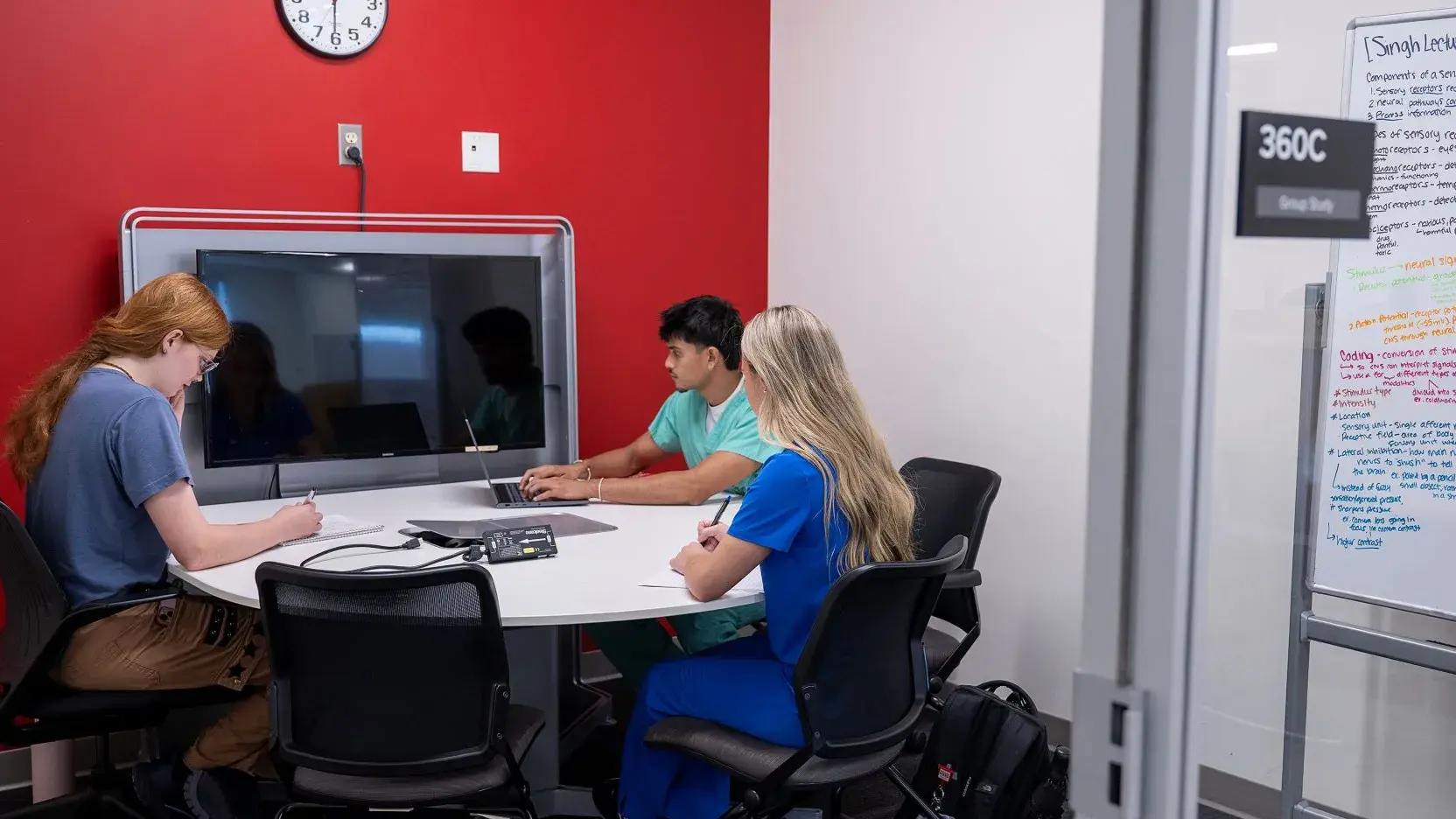 Group of students sitting around a table in the 360C study room, featuring a TV and large whiteboard.
