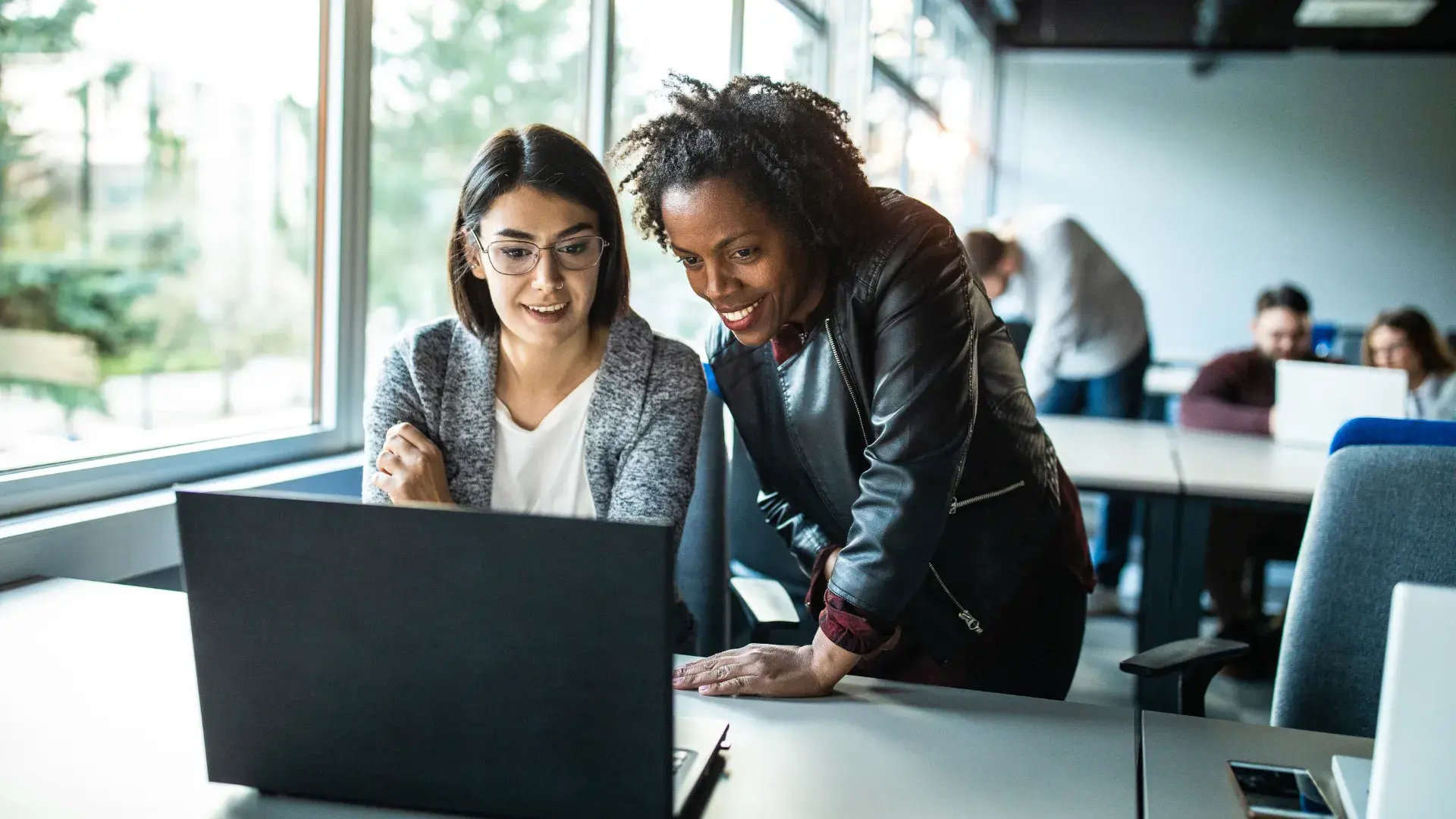 Two women collaborate at a laptop in a bright office; one sits with glasses, the other leans in to view the screen.