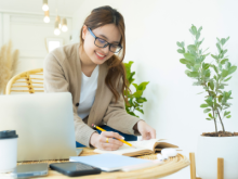 Woman sitting at a table with a laptop as she write in a notebook.